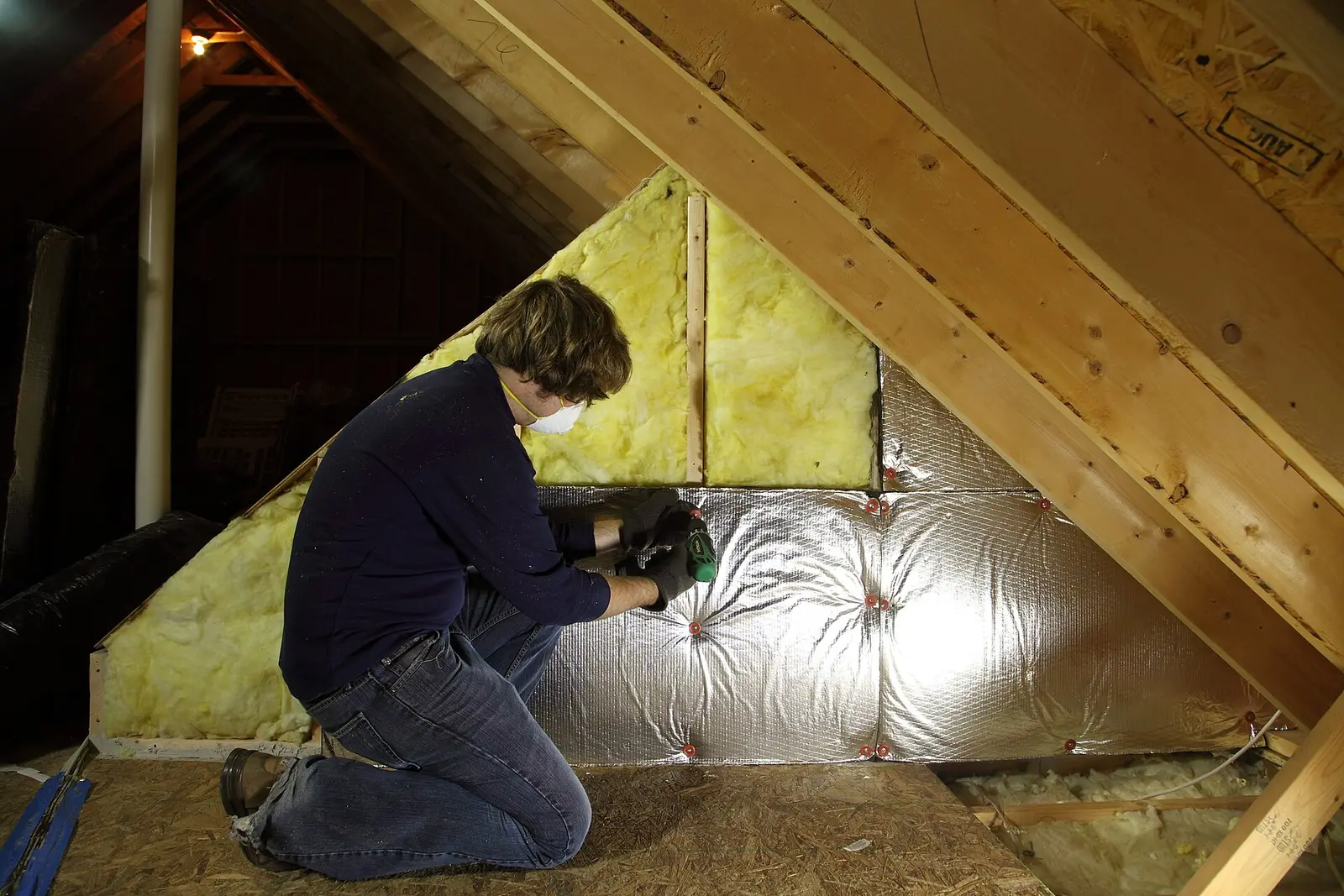 A worker in a mask fitting insulation batts between the rafters of an attic