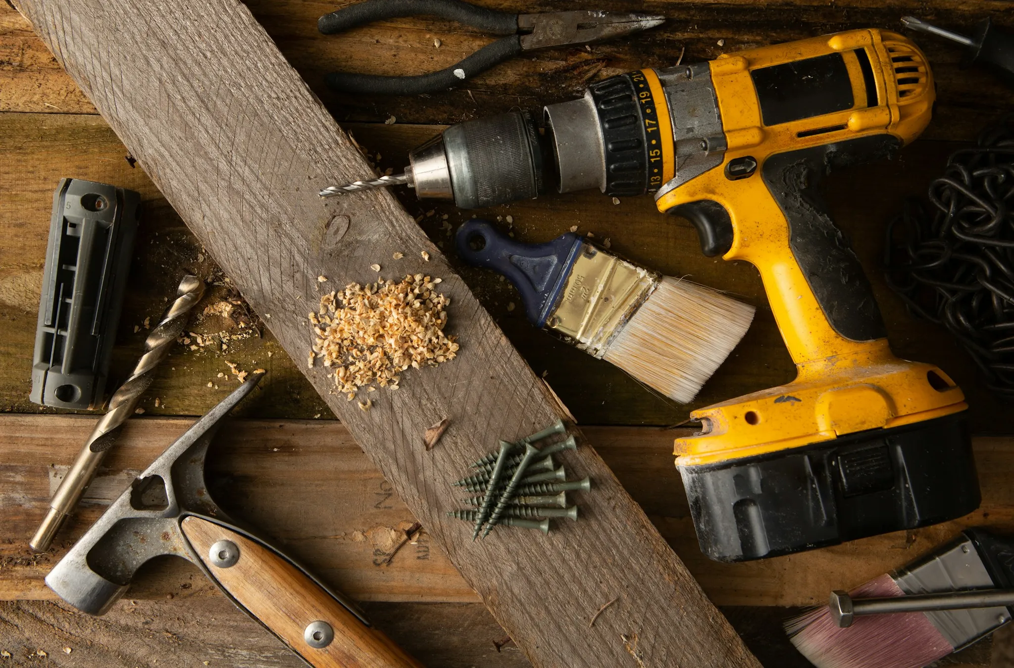 Cordless drill, hammer, paintbrush, screws and drill bits on a wooden workbench