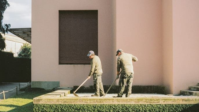 Two workers raking a raised yard area beside a pink stucco building