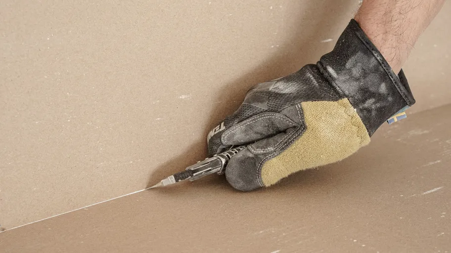 Gloved hand scoring a drywall sheet with a utility knife
